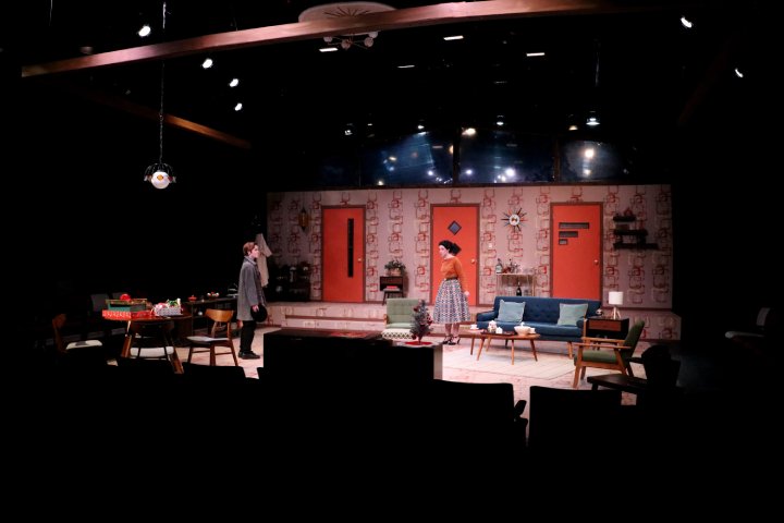 Two actors perform on a stage in a black box theater, set up as a mid-century living room with vintage furniture and decor. The lighting focuses on the actors, highlighting the retro wallpaper and orange doors in the background.